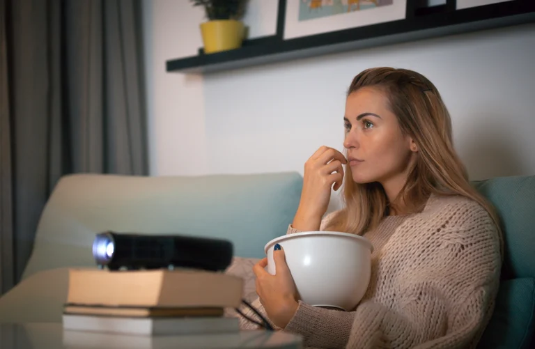 Frau auf Sofa schaut mit einem Beamer und Popcorn auf eine Leinwand